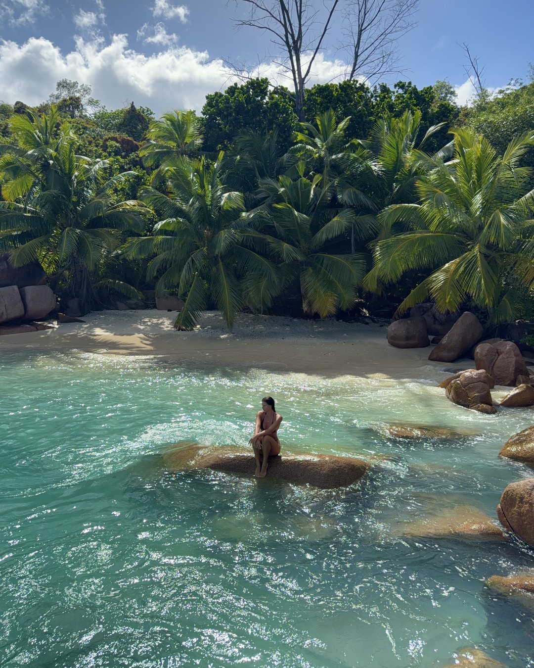 A woman sitting on a granite boulder in shallow turquoise water at the secret beach past Anse Lazio, Praslin, Seychelles
