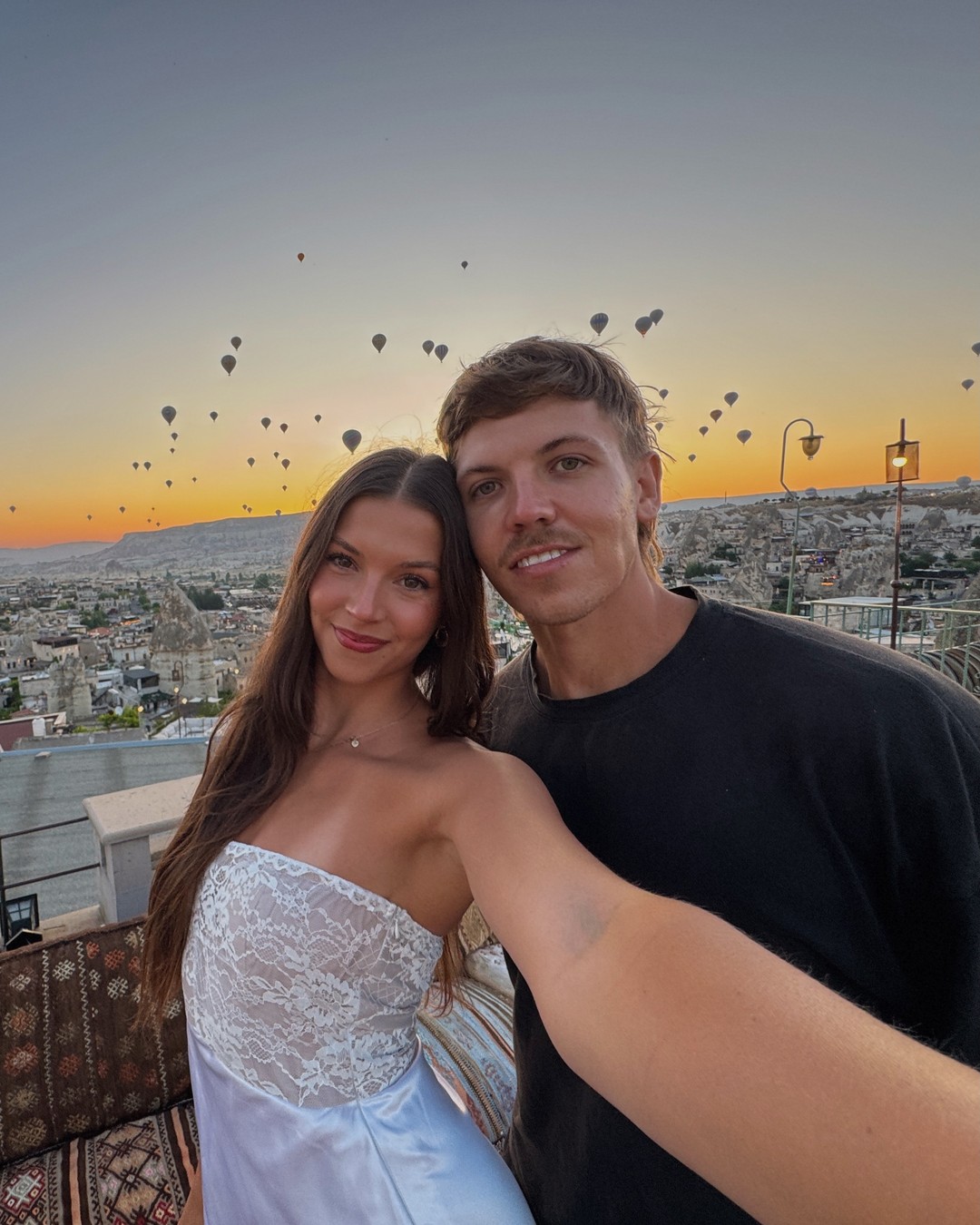 Couple on a cave hotel rooftop at sunrise with hot air balloons rising over Cappadocia